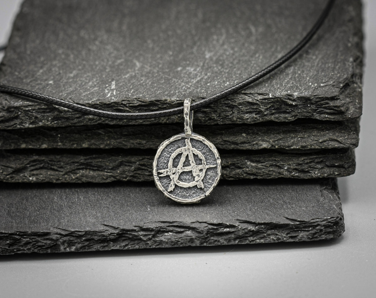 A silver-toned circular pendant necklace featuring an anarchy symbol, resting on a black cord over stacked dark slate tiles .