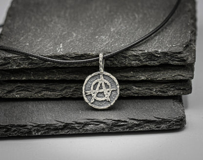 A silver-toned circular pendant necklace featuring an anarchy symbol, resting on a black cord over stacked dark slate tiles .