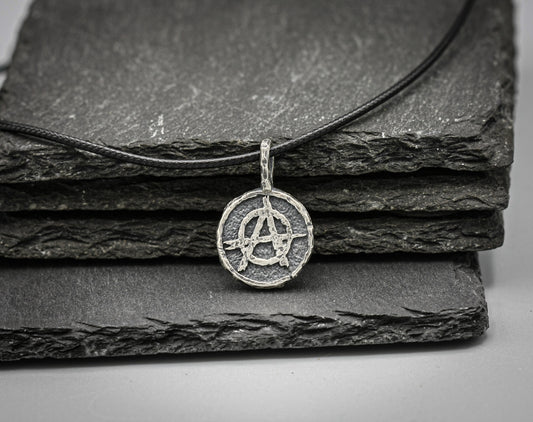 A silver-toned circular pendant necklace featuring an anarchy symbol, resting on a black cord over stacked dark slate tiles .