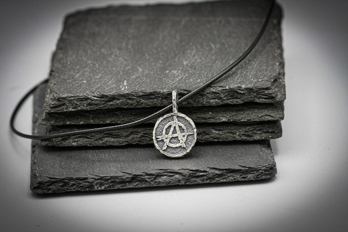 A silver pendant necklace with an anarchy symbol, resting on a stack of dark slate tiles against a light gray background.