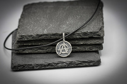 A silver pendant necklace with an anarchy symbol, resting on a stack of dark slate tiles against a light gray background.