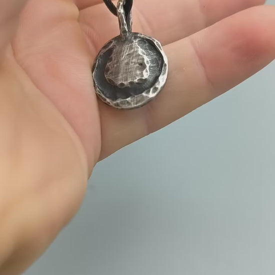 Sterling silver anarchy pendant lying flat on slate, showing raised symbol, textured finish and black cord